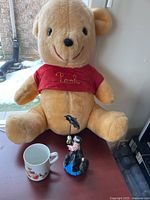Winnie the Pooh plush bear sitting behind Snow White mug and Mary Poppins figurine on table.
