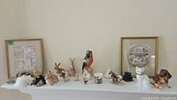 Shelf showing framed prints behind rows of various animal figurines including rabbits, birds, cats, dogs, plus a small ceramic cup and a metal wristwatch.