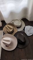 Five hats arranged on a floor: a white baseball cap, two straw hats, a dark brown wide-brimmed hat, and a beige felt hat with feathers.