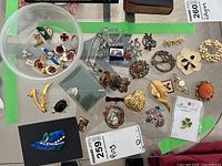 Full lot view showing variety of pins and brooches including safety pins, floral brooches, and themed pins in a small container and spread out on a surface.
