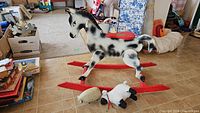 Wooden rocking horse with spotted black and white pattern and red rockers on tiled floor, accompanied by a plastic sheep and a stuffed sheep toy.
