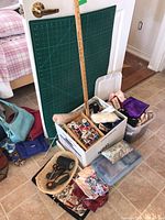 Full lot showing cutting mat, ruler, large white box with thread spools, fabrics, pincushion, and wicker basket with sewing tools on tiled floor.