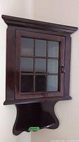 Full view of the wood corner hanging cabinet mounted on the wall, showing the glass door and the wooden shelf beneath it.