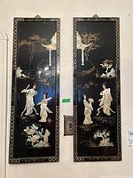 Two black lacquered rectangular panels with decorative frames, each featuring mother-of-pearl inlaid figures of women along with gold line art of natural and architectural elements.