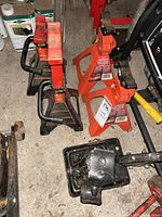 Four automotive jacks and stands; two red and black axle jacks and two orange axle stands with triangular cutouts. Shown on garage concrete floor beside other garage items.