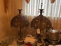 Full view of the pair of lamps showing filigree shades with hanging glass prisms, cherub bases, and decorative stone pedestals. One lamp missing finial.