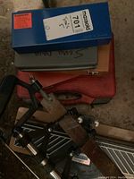 Photo shows a blue case containing hole saw drill bits, stacked on smaller gray and orange tool cases, with part of a hacksaw and other saw blades visible around.