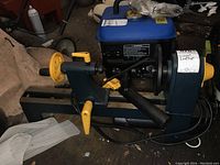 Photo of entire lathe showing cast iron bed, tool rest, yellow adjustment handles, black and blue body with motor setup behind, captured in garage environment.