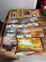 Photo showing all twelve unframed oil paintings arranged on a wooden table including winter scenes, portrait, and boats