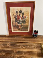 Full view of framed print with three soldiers in red jackets, tartan kilts and black fur hats standing near a barrel; Pepsi can for scale.