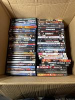 Overhead view of a cardboard box filled with two stacks of DVDs arranged vertically. Titles include 'Breach,' 'Plan B,' 'Shaft,' among others.