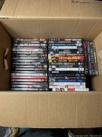 Top view of a cardboard box filled with DVDs showing their spines. Various titles primarily in action and drama genres are visible.