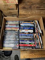 Photo showing a cardboard box filled with DVDs stacked in two rows, titles visible such as Divergence, The Departed, Stigmata, The Expendables.