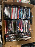 Top-down view of a box filled with two layers of assorted DVDs. Movie titles visible include Die Hard, The Mummy, The Company You Keep and John Wick.