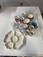 Overview of all kitchen glassware and mugs on a white table showing the assortment of items including glass tumblers, mugs, and a sectioned serving dish.