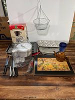 Overview photo of all kitchen items displayed on wooden surface including knives, coffee dripper and filters, hanging metal basket, baking pans, trays, teapot, and kitchen tools.