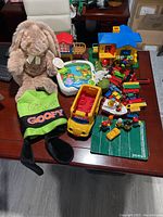 Full lot of mixed kids toys including plush bunny, Goofy jacket, toy vehicles, Fisher-Price game, and toy houses arranged on table