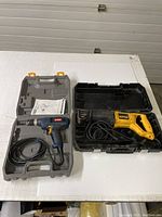 Ryobi drill in case on the left, DeWalt reciprocating saw in case on the right on white table.