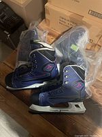 Two pairs of navy blue ice skates in plastic bags on wooden floor with cardboard boxes in background.