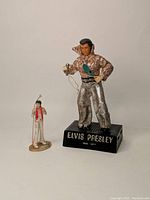 Photo showing both Elvis Presley figurines side-by-side, one larger in shiny silver and gold on a black base, the other smaller in a white jumpsuit with a red scarf on a beige base.
