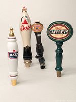 Four vintage beer tap handles shown in profile, aligned side-by-side including Harp, Bass, Stella Artois, and Thomas Caffrey's