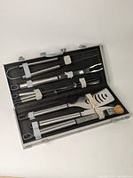 Opened view showing BBQ utensils inside case, held by elastic bands, including spatula, fork, tongs, and basting brush.