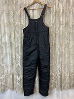 Full front view of black snowpants hanging on a wooden hanger, showing front zipper and overall design.