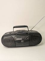 Front view of black RCA portable stereo with two speakers flanking a cassette player and control buttons, antenna extended from back right side.