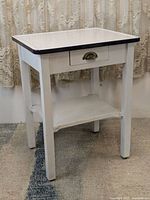 Front angle of solid wood farmhouse table showing white enamel top, drawer with brass pull, and lower shelf.