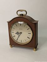 Front view of Junghans Electro-Gong clock showing wooden case, brass handle, brass feet, closed glass door, and clock face with Roman numerals and ornate clock hands.