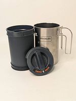 Disassembled parts of the Stanley stainless steel coffee press: main body, detachable plunger inside black container, and black lid with marked text.