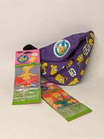Purple fabric Simpsons fanny pack with character faces and quotes, showing an emblem with Simpsons character, photographed folded on a white background.
