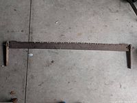 Photo of traditional antique cross cut saw with long serrated blade and wooden handles on a concrete floor.