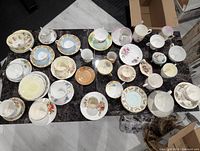 Full overview of assorted cups and saucers laid out on black surface showing various floral patterns and design styles.