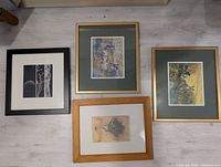Four framed prints laid flat on a light floor. Two prints have gold frames with green mats, one has a black frame and one a light wooden frame.