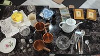 Full view of assorted items including glassware, copper mugs, silverware, teapot, framed art, and more on a dark marble surface.