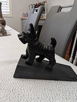 Black cast iron Yorkie dog doorstop shown on a white textured tablecloth with dining chairs in the background.