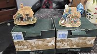 Two Lilliput Lane miniature cottages on top of their original green boxes, showing the front and side with boxed packaging and village illustrations.