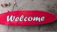 Top view of the red wooden welcome sign with white cursive lettering on a cement floor with a few leaves around it.