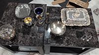 Collection of silver and silver-tone bowls, cups, lidded containers, and decorative tray spread out on two black marble tables.