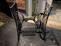 Full view of Singer sewing machine treadle base on driveway, showing the base structure and gold 'SINGER' lettering on horizontal bar