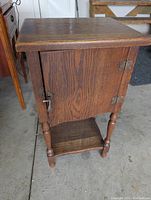View of the older wooden cabinet with closed door showing metal hinges and latch, turned legs, and shelf at bottom.
