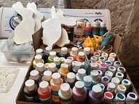 Box containing approximately 50+ bottles of acrylic paint in assorted colors, various brands mainly Apple Barrel and DecoArt, with 6 wooden rabbit cut outs behind the box along with small decorative items shaped as cheese and birds