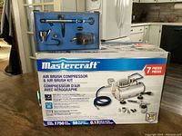 Mastercraft airbrush compressor in original box alongside blue case containing airbrush spray gun and accessories.