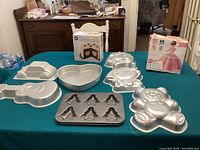 Photo showing seven assorted character cake pans on table, including teddy bear, guitar, car, heart, flower, and gray non-stick mini angel fairy pan, along with boxed doll cake mold.