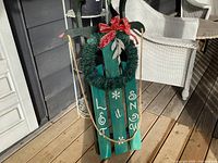 Green wooden sled with metal runners on a wooden porch floor, decorated with a green wreath and red bow. White lettering says 'Let it Snow' with snowflake accents.