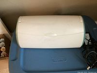 White closed Cricut cutting machine on blue storage bin with power cord visible.