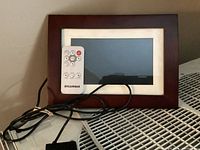 Photo of Panasonic digital photo frame with white bezel and dark wood frame, including Sylvania remote control and power cord.