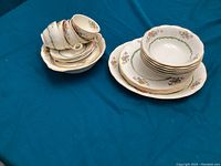 Overall view of antique Grindley dinnerware set stacked on teal tablecloth, showing cups, saucers, small bowls, and plates with floral and green garland pattern