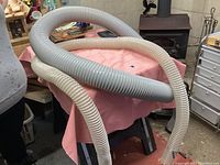Two flexible dust collector tubes coiled on a pink cloth-covered stand. One tube is gray with larger 6 inch diameter, the other is white with smaller 4 inch diameter. Both tubes ribbed and designed for dust collection in woodworking.